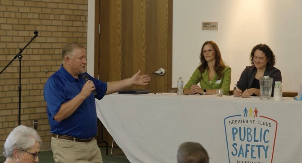 A man in a blue shirt speaks into a microphone while gesturing with his right hand. He stands in front of a seated audience. Two women sit at a table with a sign that reads "Greater St. Cloud Public Safety Foundation," listening attentively.