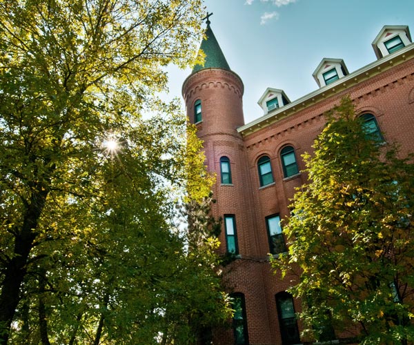 A tall, red-brick building with round turret and conical roof stands amidst lush green trees. The sun filters through the tree leaves, casting dappled light on the building's windows. The sky is clear and blue.