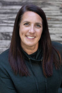 A woman with long brown hair is smiling, wearing a dark green sweater. She is sitting outdoors on a wooden surface with a blurred background.