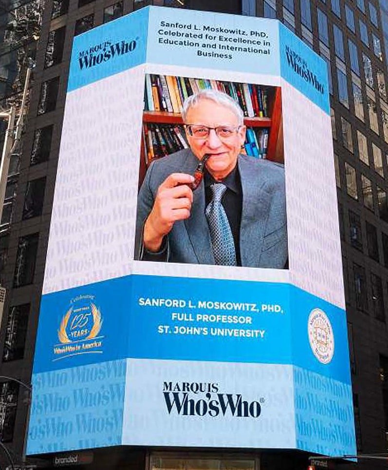 A large digital billboard in Times Square features an older man sitting with a bookcase in the background. He holds a pipe. Text around him highlights his achievements and titles, including his name and recognition for excellence in education and business.