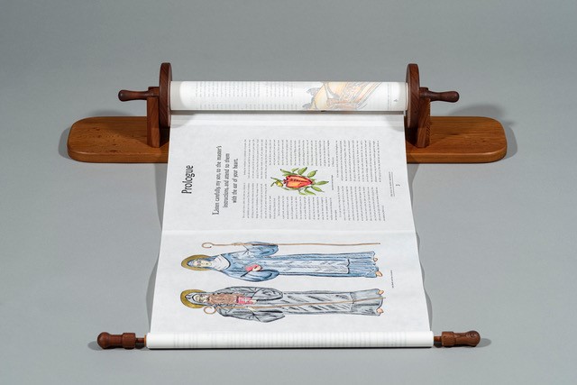 A scroll displayed on wooden holders, showing a page titled "Prologue." The page features illustrations of two robed figures and a decorative heart symbol. The scroll is partially unrolled on a gray surface.