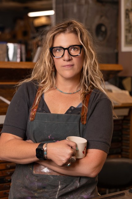 A person with long hair and glasses wearing a dark shirt and apron, holding a mug. They stand in a workshop environment, with shelves and supplies in the background, creating a cozy and creative atmosphere.