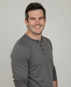 A man with short, dark hair is smiling and wearing a gray long-sleeve henley shirt. He is standing against a light, neutral background, with his body slightly angled to the side.