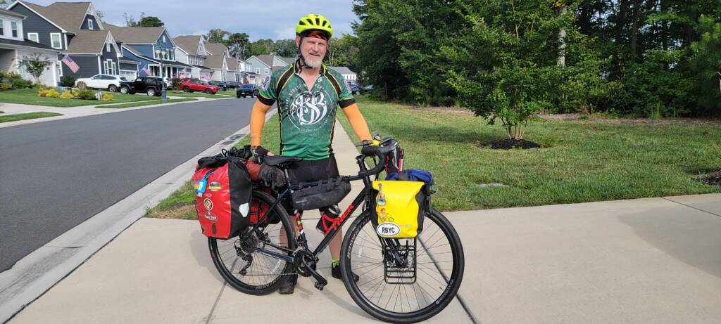 A cyclist in a green jersey and yellow helmet stands with a touring bike loaded with red and yellow panniers in a suburban neighborhood. Houses and trees line the street in the background.