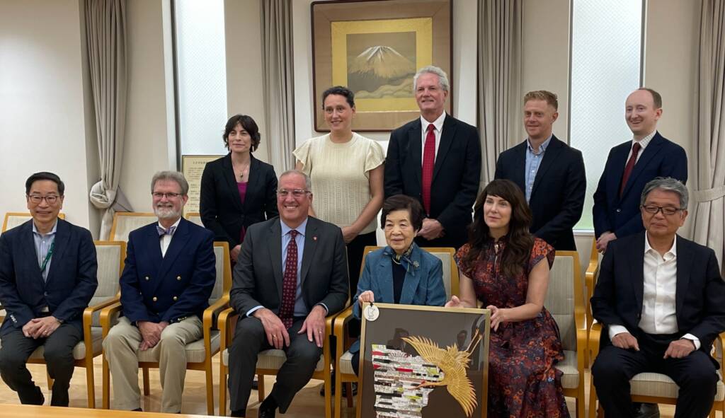 A group of eleven people are posing together in a formal setting. Some are seated while others stand behind them. One woman in the front holds a framed item with a gold bird design. A framed painting of a mountain is on the wall behind them.