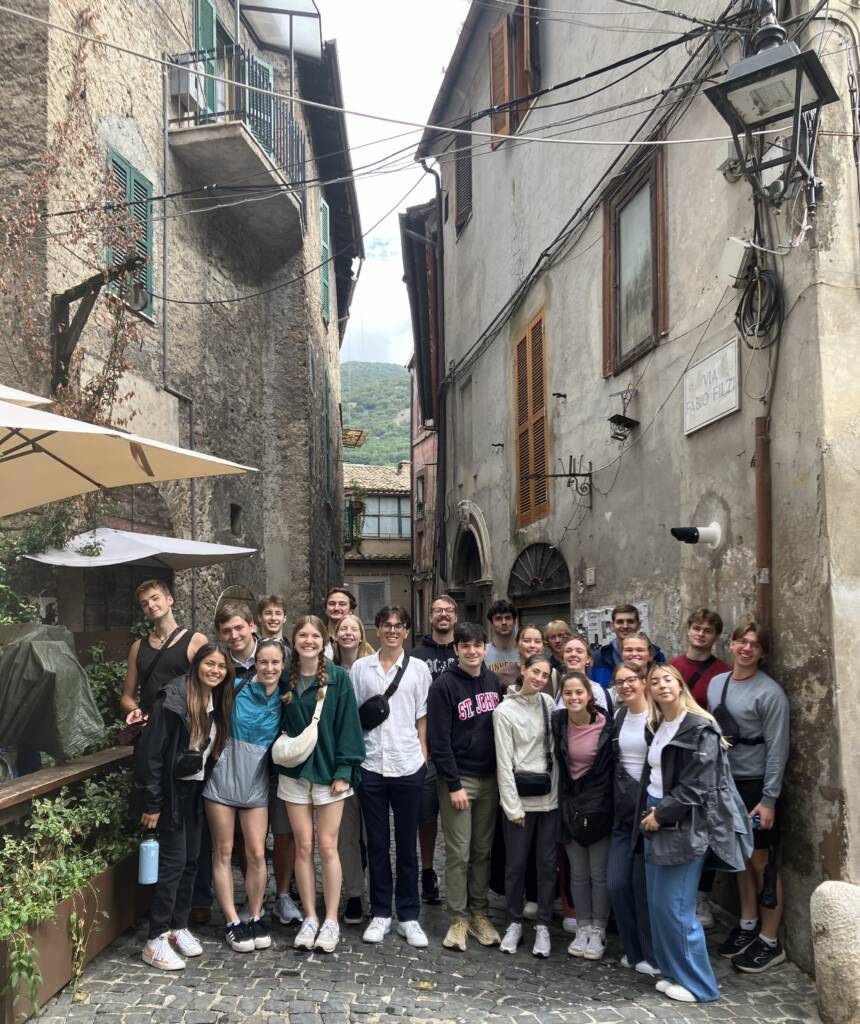 A group of people posing together in a narrow, cobbled street between old stone buildings. Some wear jackets and hold umbrellas. The setting appears to be a historic European town, with plants and narrow balconies visible.