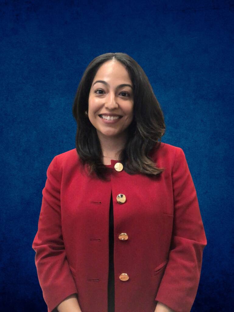 A person with long dark hair is smiling, wearing a red jacket with gold buttons, in front of a blue background.