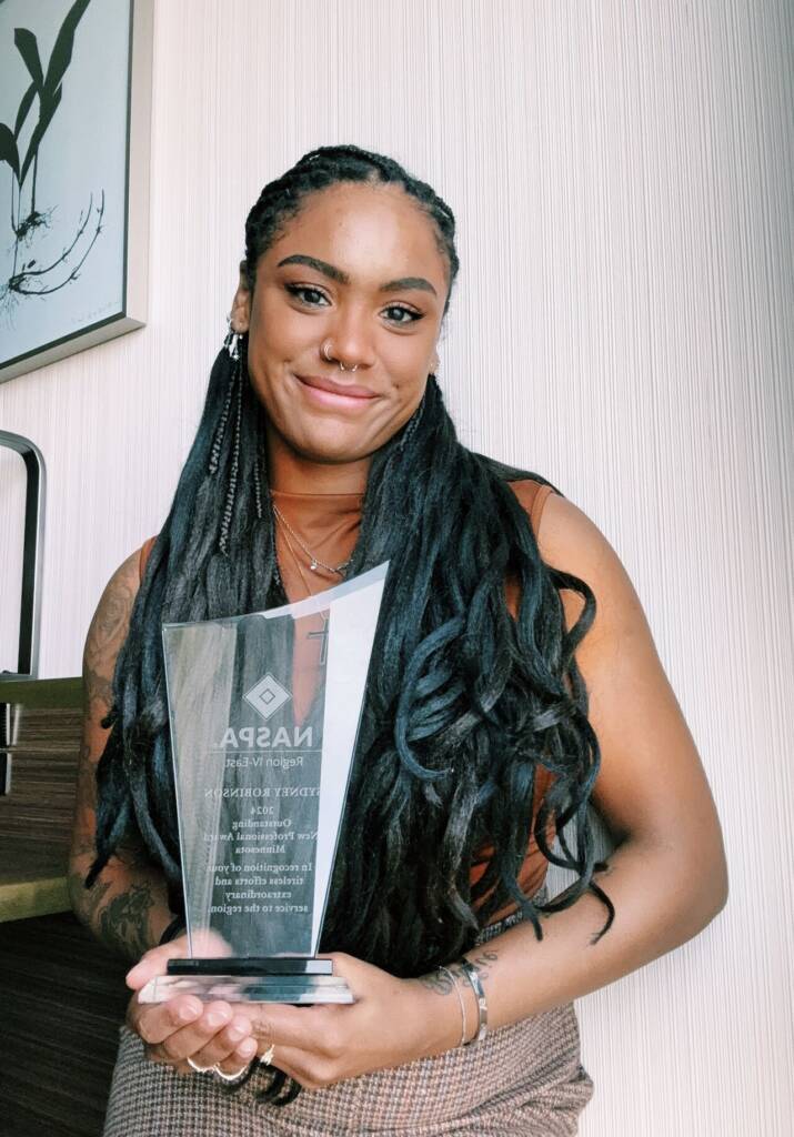 A person with long braided hair smiles while holding an award. They are standing in front of a textured wall and wearing a sleeveless top. The award has text on it, though it is not fully legible in the image.