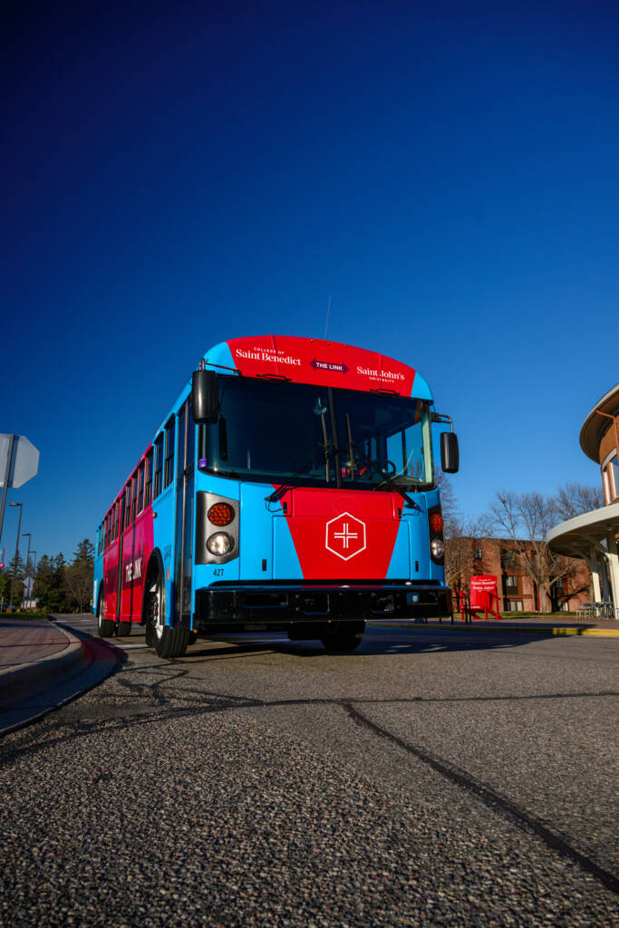 A red and blue bus with a shield symbol on the front is parked on a street under a clear blue sky. Buildings and trees are visible in the background.