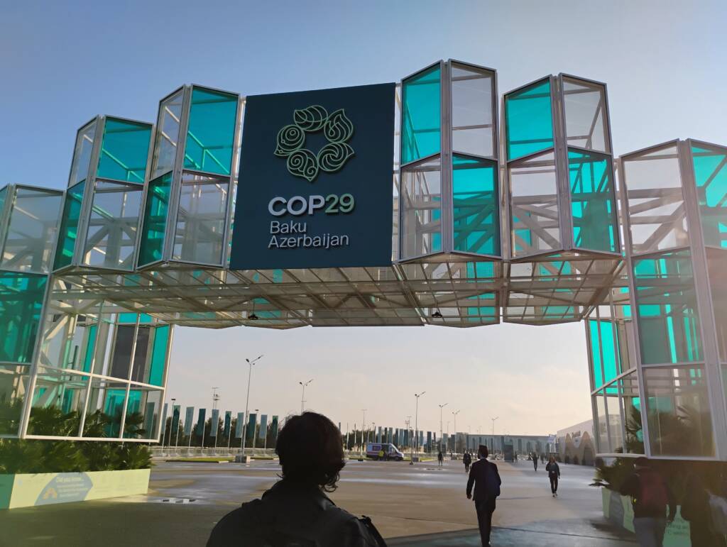 A person walks towards a large, glass-paneled structure displaying a sign for COP29 in Baku, Azerbaijan. The structure features a teal and white design and the sky is clear in the background.