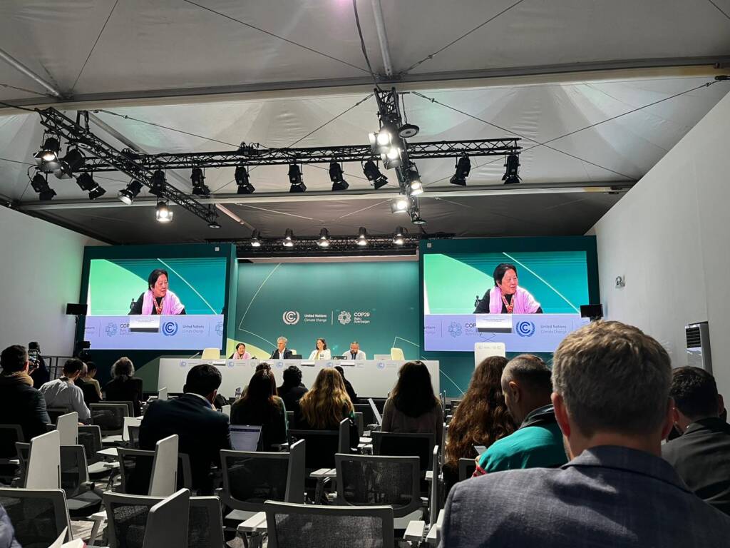 A conference room with a speaker on stage, visible on large screens. Attendees sit facing the stage. The setting is formal, with a green and white backdrop displaying logos and text. Ceiling lights illuminate the room.