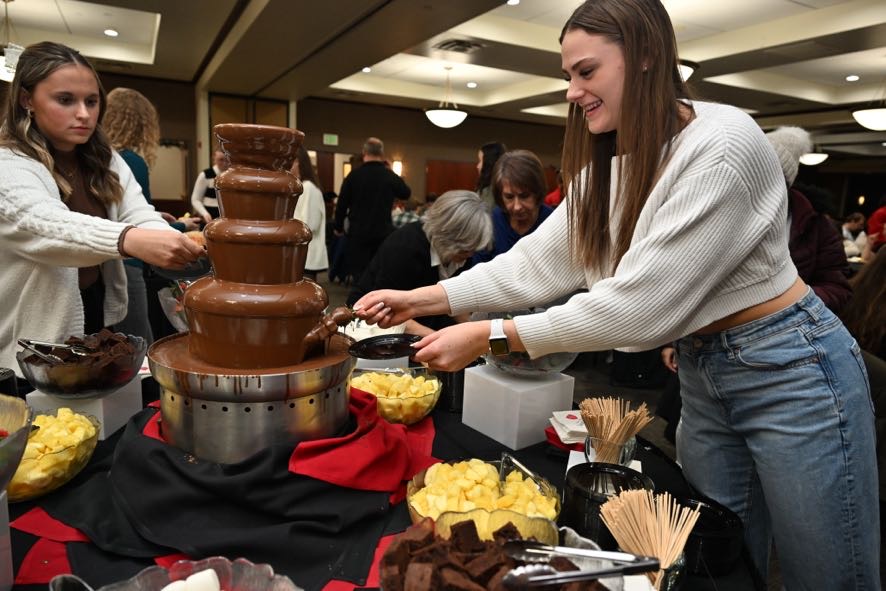 A woman in a white sweater smiles as she dips food into a large chocolate fountain at a buffet table filled with pineapple chunks, brownies, and skewers. Other people are gathered around, enjoying the event in a warmly lit room.