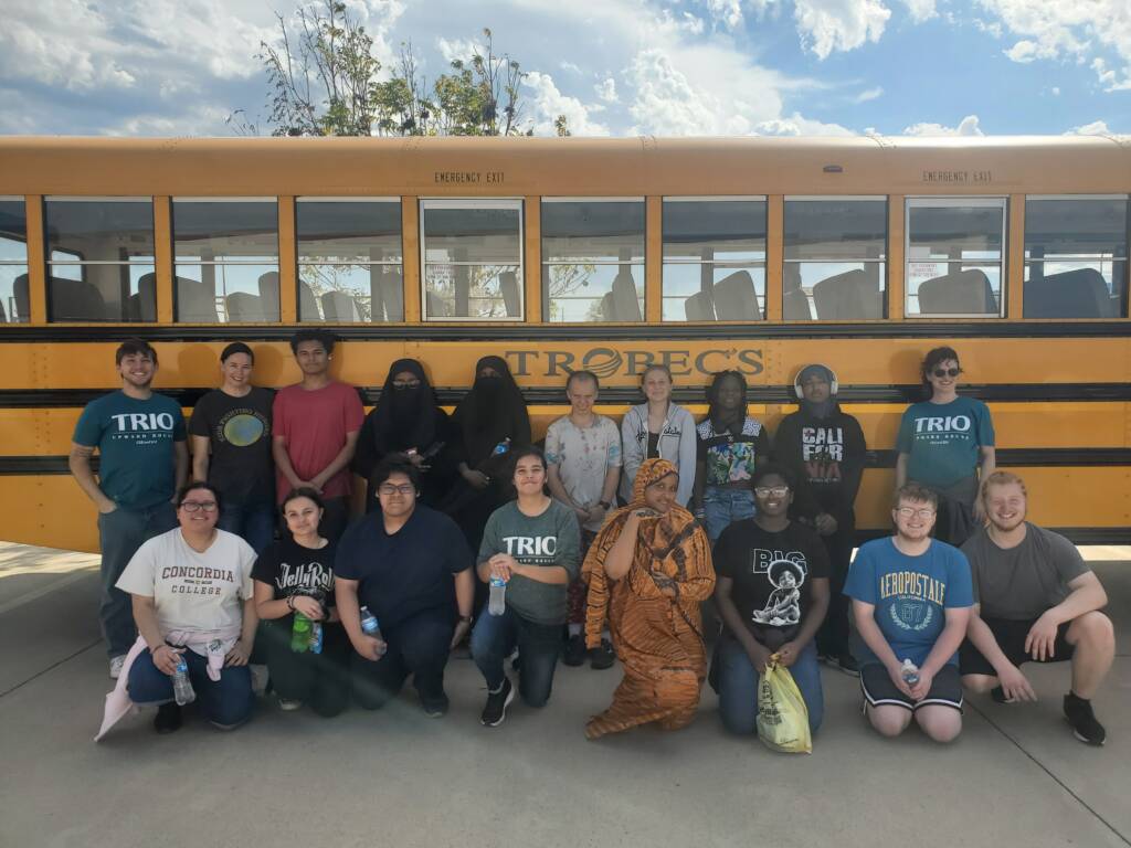 A diverse group of people posing in front of a yellow school bus. Some are seated while others stand; they appear happy. The sky is partly cloudy, and there are a few trees in the background.