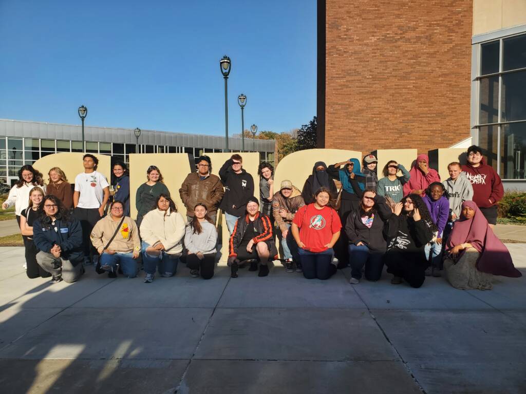 A group of people posing together outside a building with a large "STATE" sign. Some are sitting while others stand behind them. They are dressed in casual attire and are smiling under a clear blue sky.