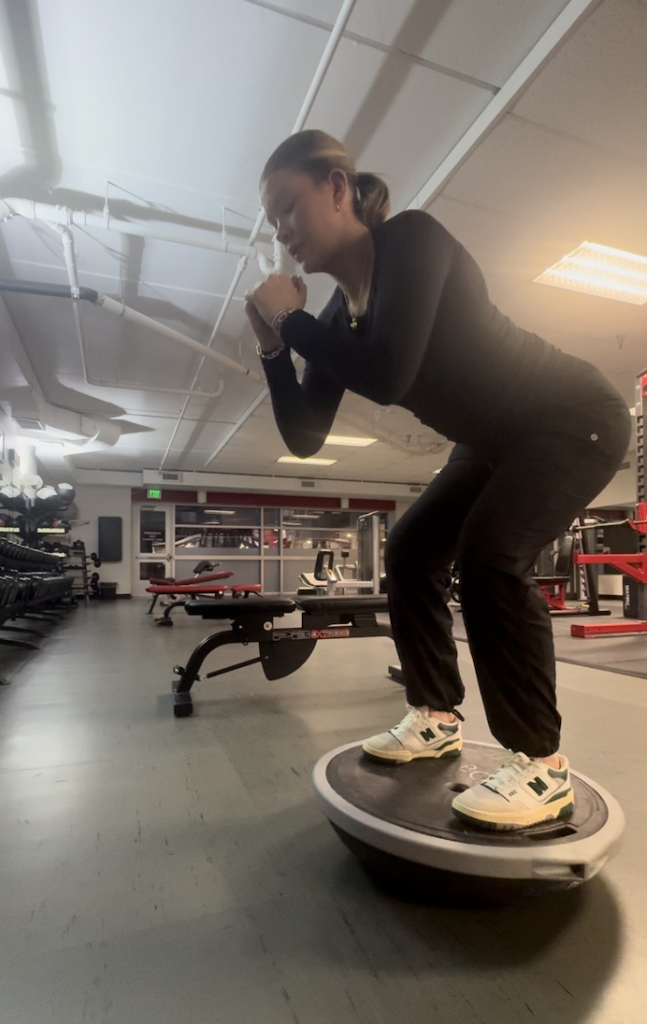 A person balancing on a BOSU ball while performing a squat in a gym setting. The individual is wearing black workout clothes and white sneakers. Gym equipment is visible in the background, including benches and weights.