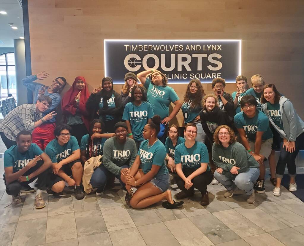 A diverse group of people, all wearing matching "TRIO" shirts, pose enthusiastically in front of a sign that reads "Timberwolves and Lynx Courts at Clinic Square." Some are kneeling or sitting in the front, others stand behind them, smiling and waving.