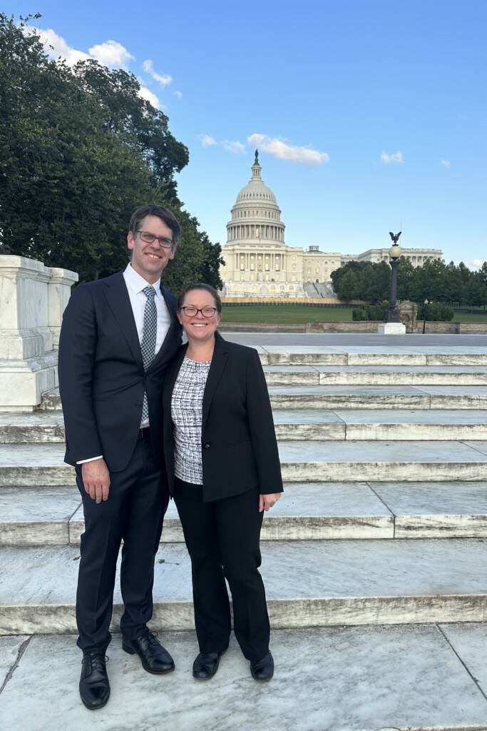 Two people stand smiling on marble steps, both dressed in business attire. Behind them is the U.S. Capitol building, partially surrounded by trees and set against a clear blue sky.