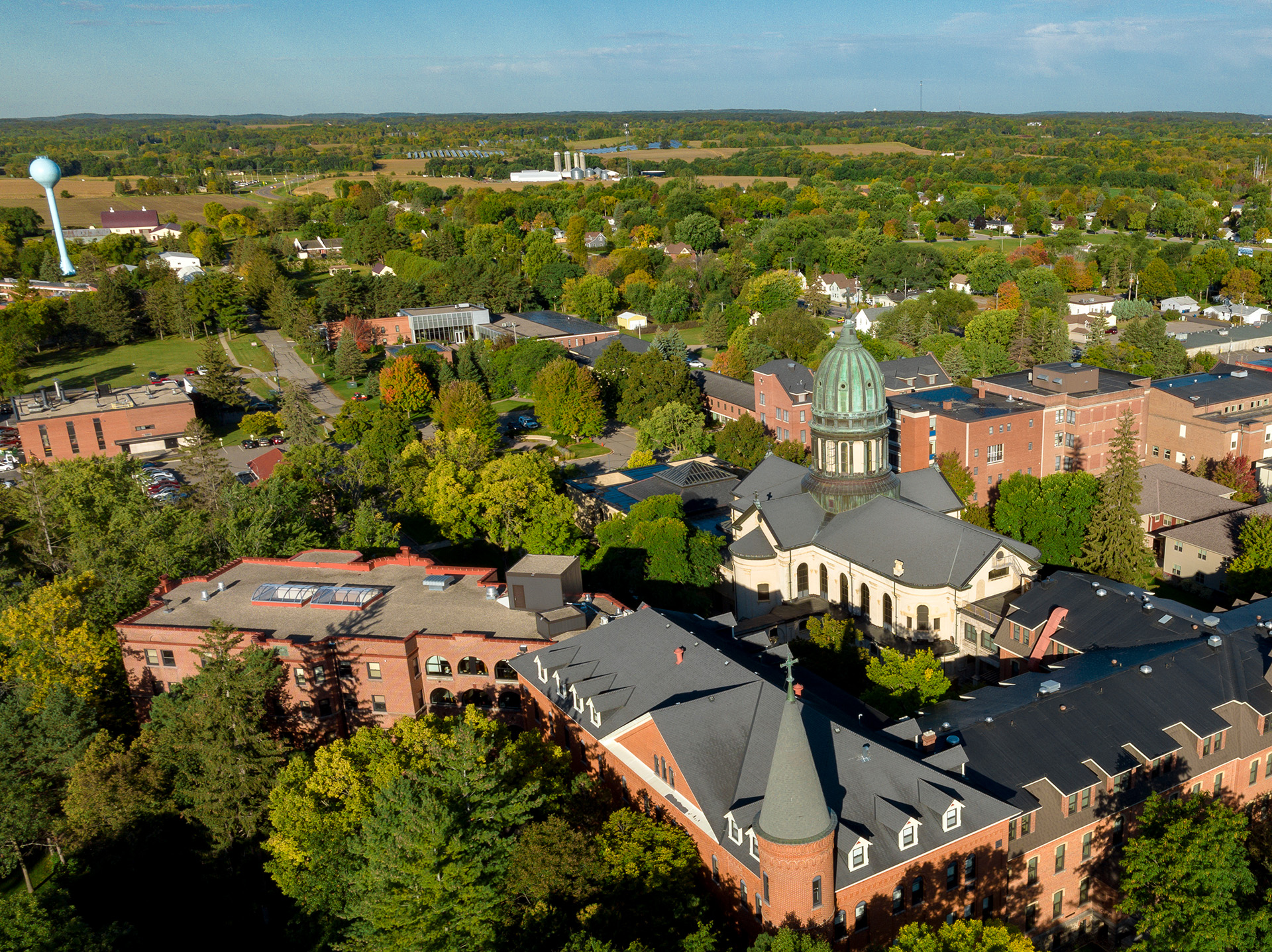 Aerial view of a campus with a prominent building featuring a domed roof surrounded by trees. Several other brick buildings are scattered throughout the area, set against a backdrop of green fields and a distant water tower under a clear blue sky.