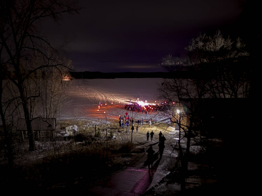 Groups of people are gathered on a snowy, illuminated path at night near a frozen lake. Trees and shadows frame the scene, and a distant light glows on the horizon.