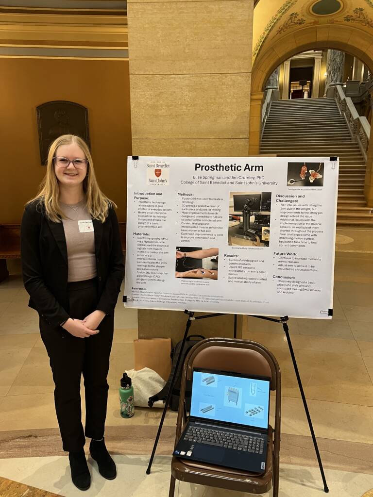 A person stands beside a poster presentation titled "Prosthetic Arm." The setup includes a laptop on a chair, displaying related information. The setting appears to be a public building with a staircase and ornate architectural details in the background.