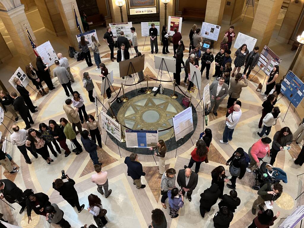 A large group of people gather in a circular lobby with an ornate star-patterned floor. They are viewing and discussing various posters displayed on easels. The scene is lively, with individuals engaged in conversations and presentations.